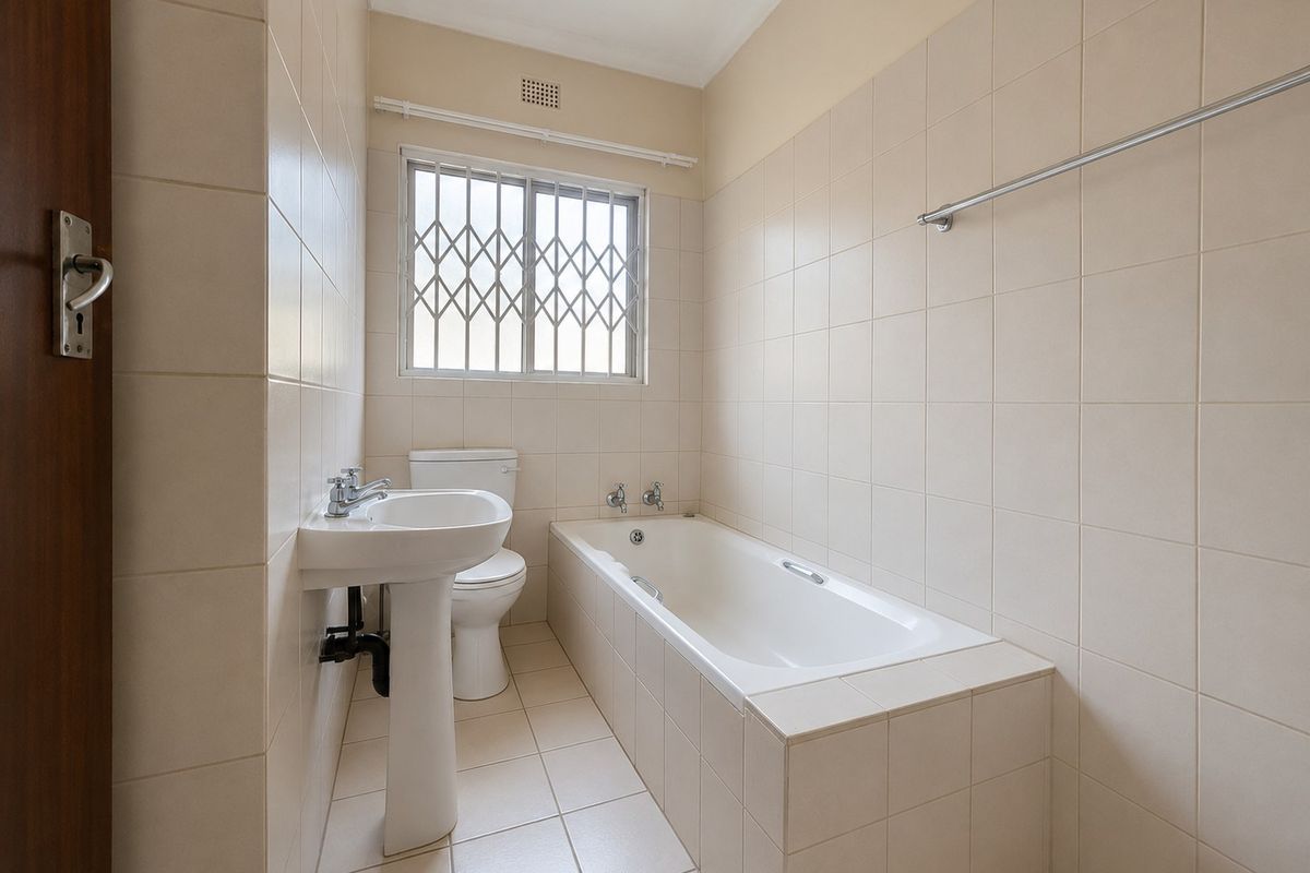 A clean, functional bathroom featuring a pedestal basin, a built-in bathtub, and neutral wall and floor tiling with a secure window.