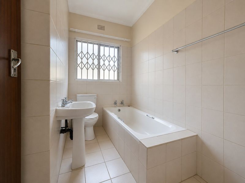 A clean, functional bathroom featuring a pedestal basin, a built-in bathtub, and neutral wall and floor tiling with a secure window.