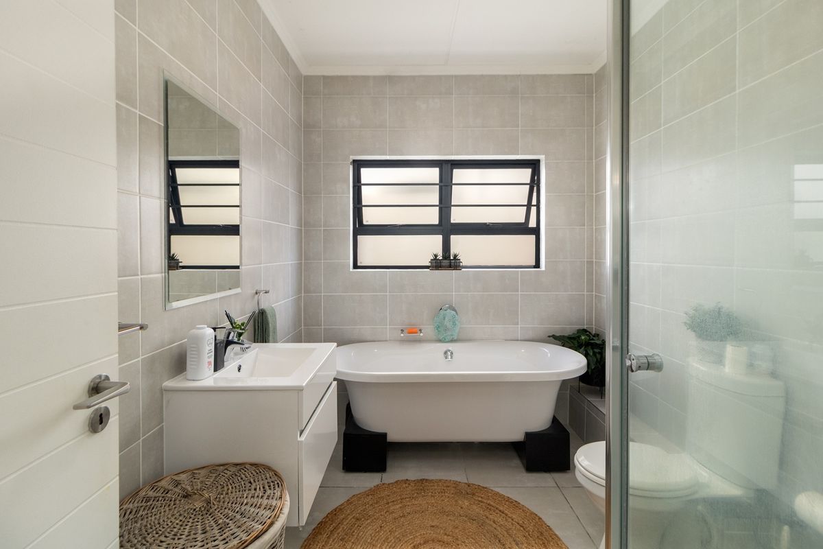 Modern bathroom featuring a freestanding bathtub, vanity with basin, glass-enclosed shower, and neutral wall tiling with a louvered window for natural light.
