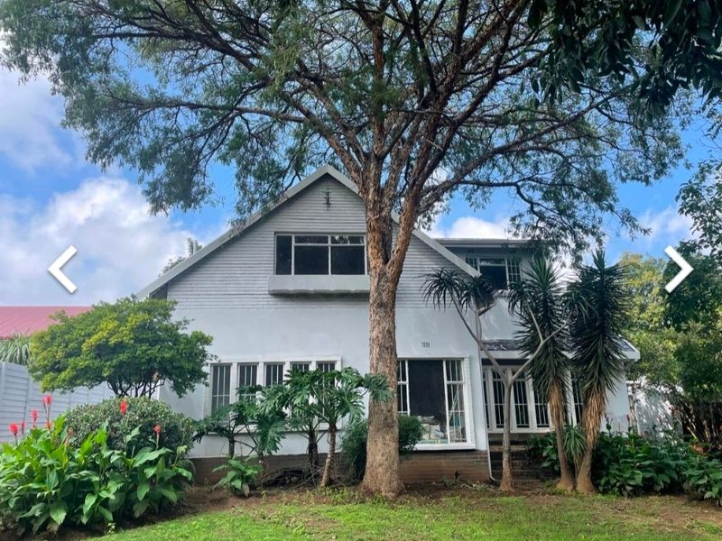 Front elevation of a two-story house featuring a pitched roof, large casement windows, and a mature garden setting.