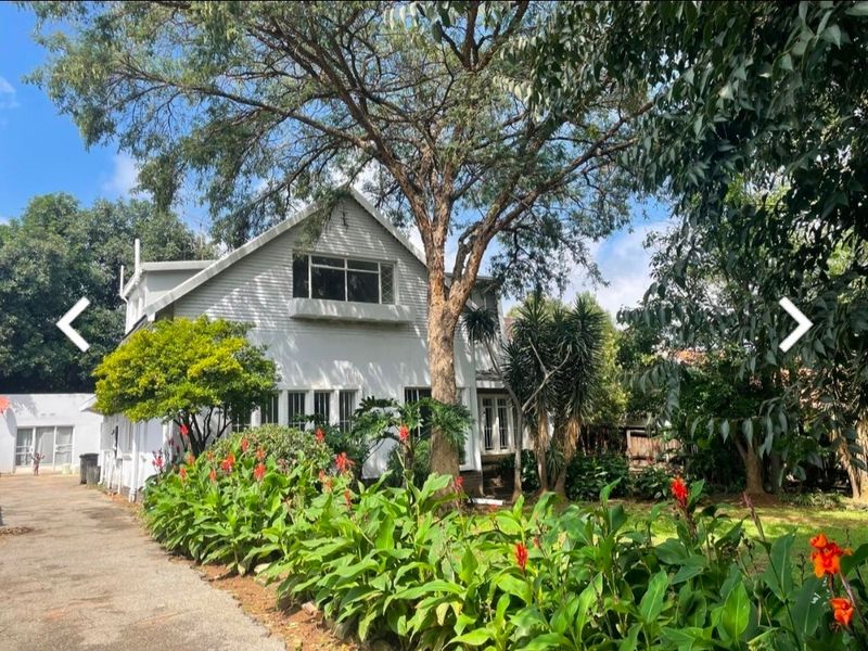 Front elevation of a two-story house featuring a pitched roof, casement windows, and a mature landscaped garden with an adjacent driveway.
