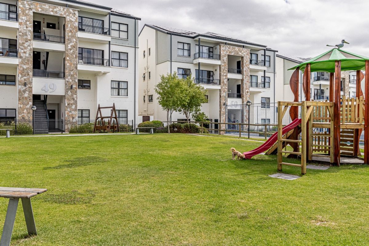 A well-maintained communal garden area featuring a playground and landscaped lawn, situated between modern apartment blocks with stone-clad architectural details.