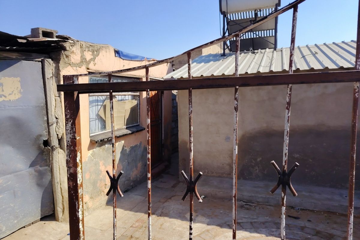 Rear exterior view of the property featuring a paved courtyard, solar water heater, and metal security gate with visible wear to the building finishes.