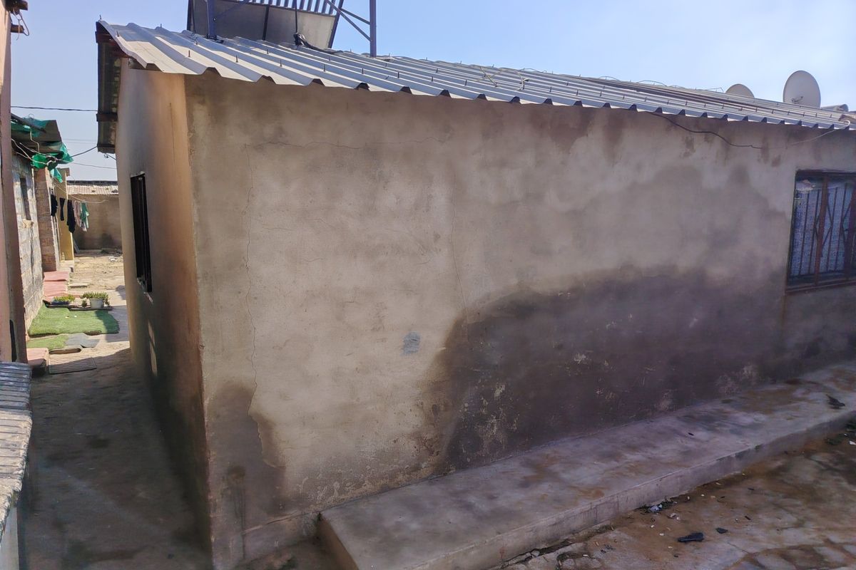 Rear exterior view of a residential structure featuring a corrugated metal roof, solar water heater, and visible signs of plaster cracking and damp staining.