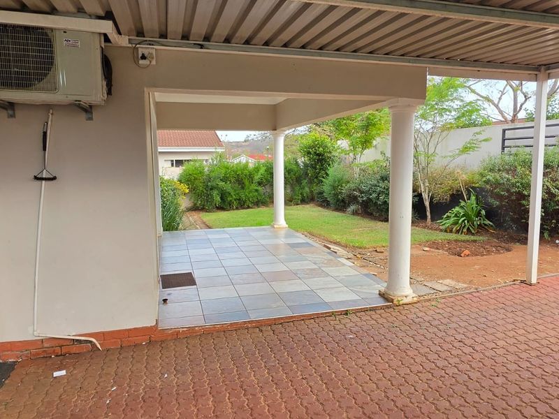 A covered patio area featuring tiled flooring and support columns, overlooking a private garden space with paved driveway access.
