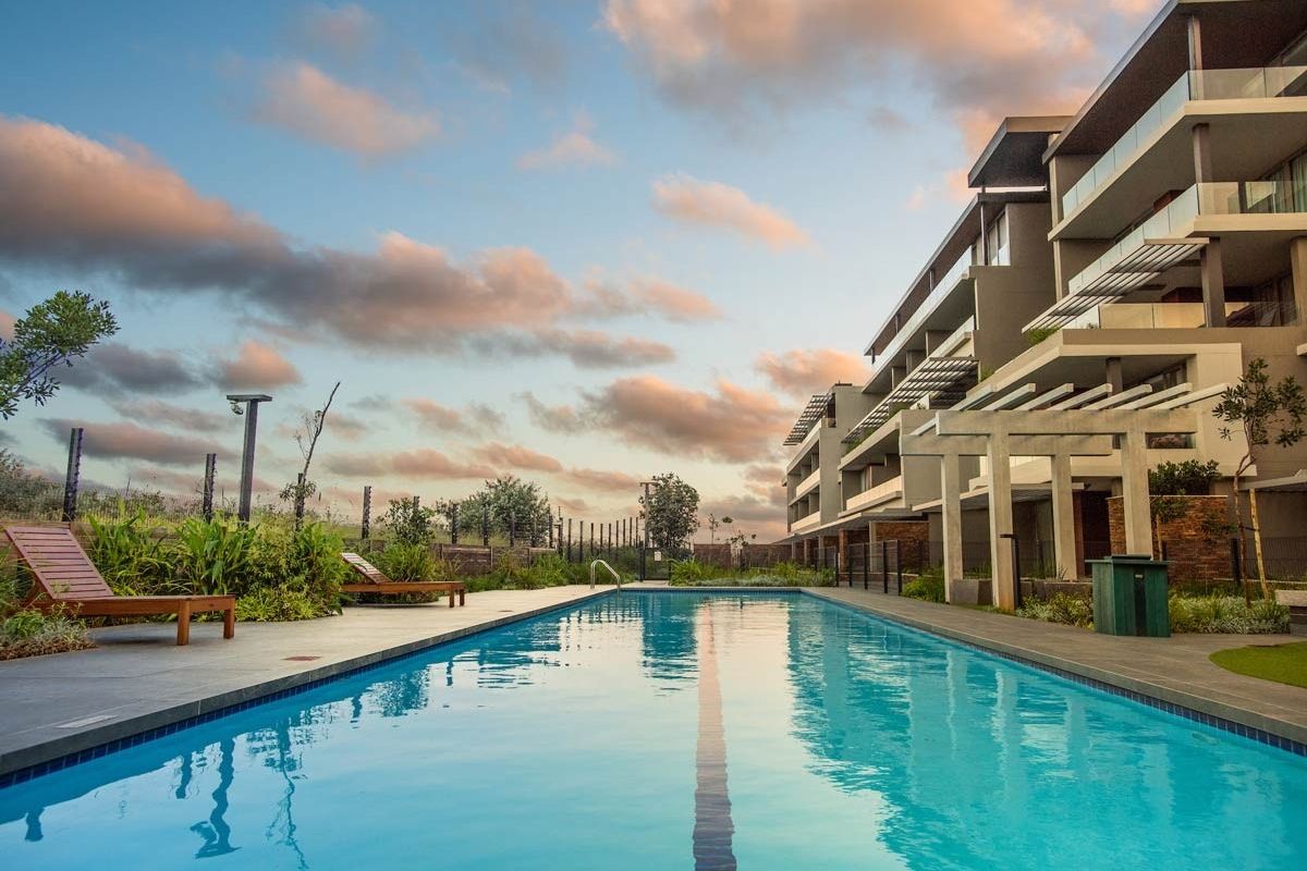 A well-maintained communal swimming pool area featuring stone paving, a pergola, and modern apartment building architecture in the background.