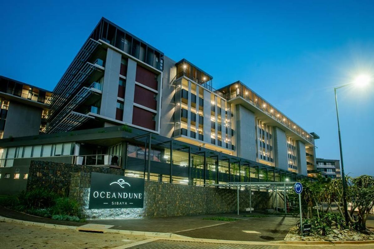 Modern multi-story apartment complex exterior featuring contemporary architectural design, stone cladding, and illuminated signage at the entrance.