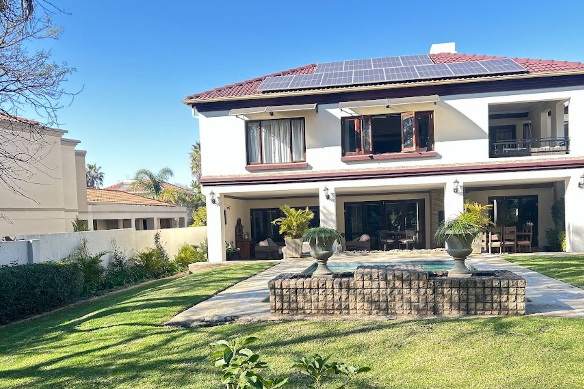 Rear view of a two-story house featuring a swimming pool, stone water feature, covered patio, and roof-mounted solar panels set within a landscaped garden.