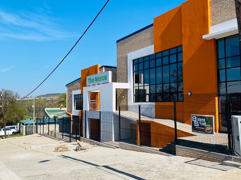 Modern office building exterior featuring a paved driveway, secure perimeter fencing, and a prominent glass facade with contemporary orange and brick architectural accents.