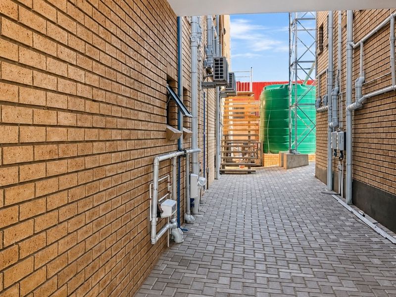 A paved exterior side passage featuring brick walls, exposed utility infrastructure, and a water storage tank at the rear.