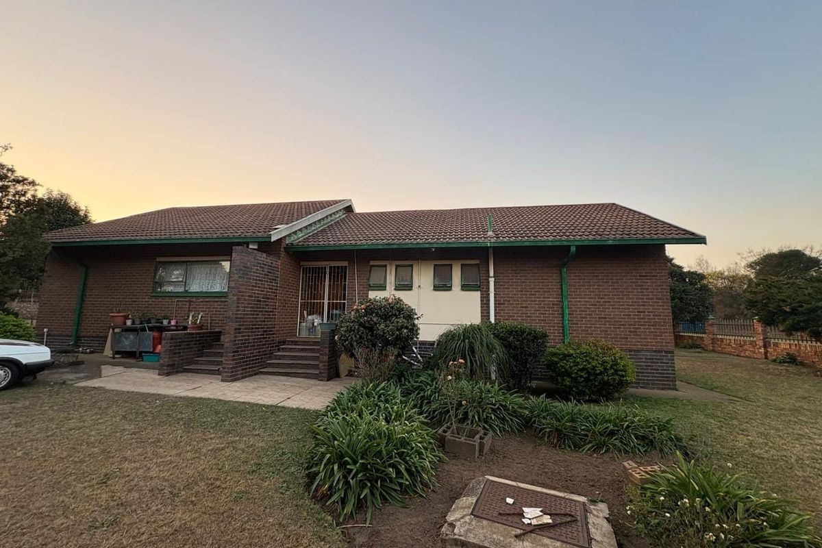 Rear view of a single-story brick house featuring a tiled roof, concrete patio area, and established garden beds.