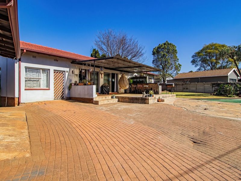 Rear exterior view featuring a paved driveway, covered patio with built-in braai, and a swimming pool in a landscaped garden setting.