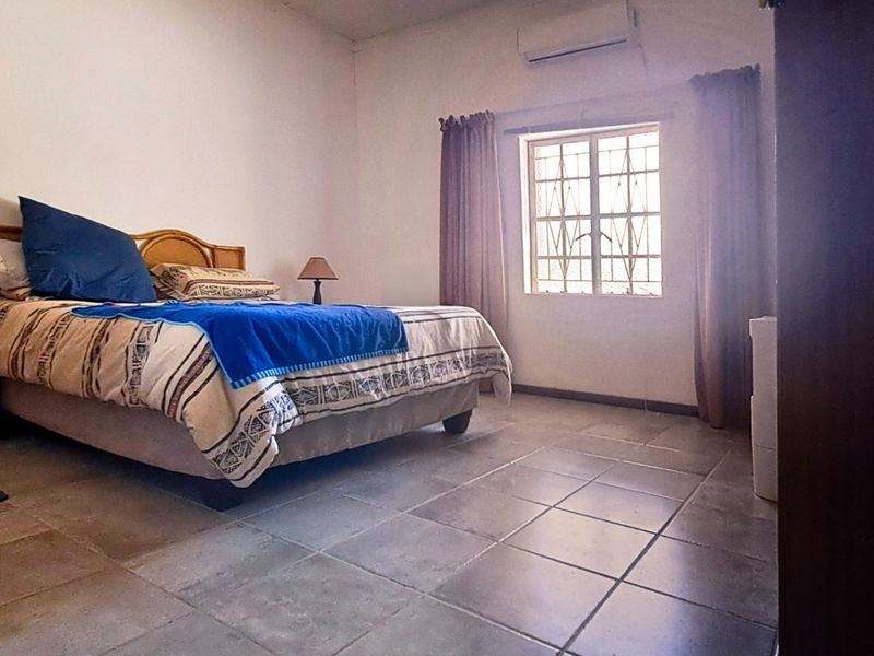 A spacious bedroom featuring tiled flooring, a wall-mounted air conditioning unit, and a window fitted with security bars.