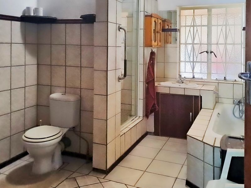 A dated bathroom featuring a built-in bathtub, shower enclosure, and vanity, with tiled finishes throughout and a security gate on the window.