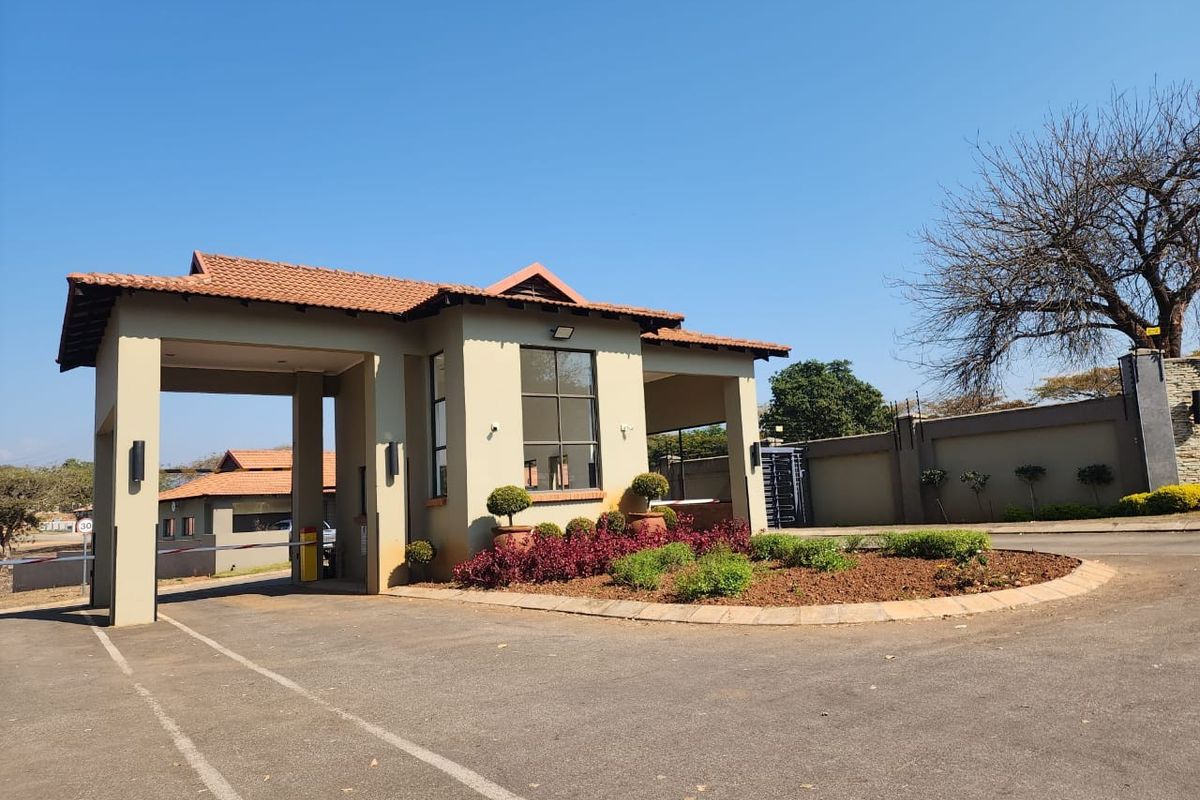 A well-maintained residential estate security gatehouse featuring a tiled roof, paved driveway, and landscaped garden beds at the entrance.