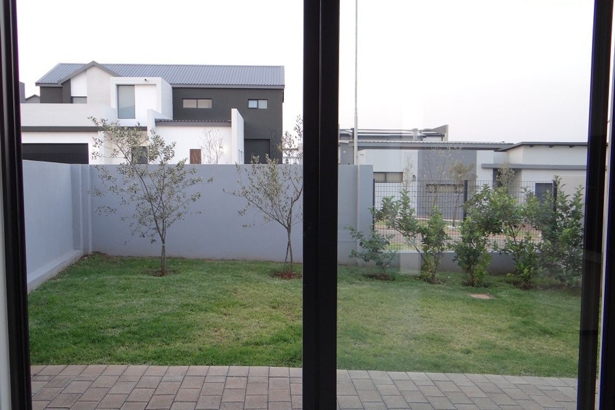 A view of a private rear garden featuring a paved patio area, manicured lawn, and perimeter boundary wall, viewed through a sliding glass door.
