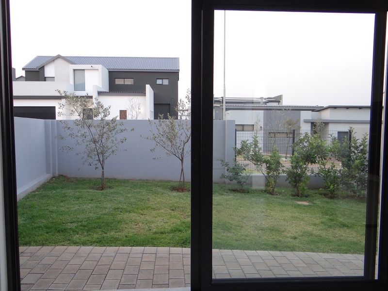 A view of a private rear garden featuring a paved patio area, manicured lawn, and perimeter boundary wall, viewed through a sliding glass door.