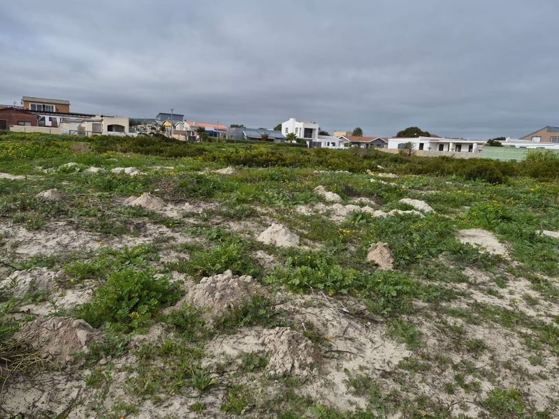 A view of a vacant residential plot featuring sandy terrain and natural vegetation, situated in a developed neighborhood.