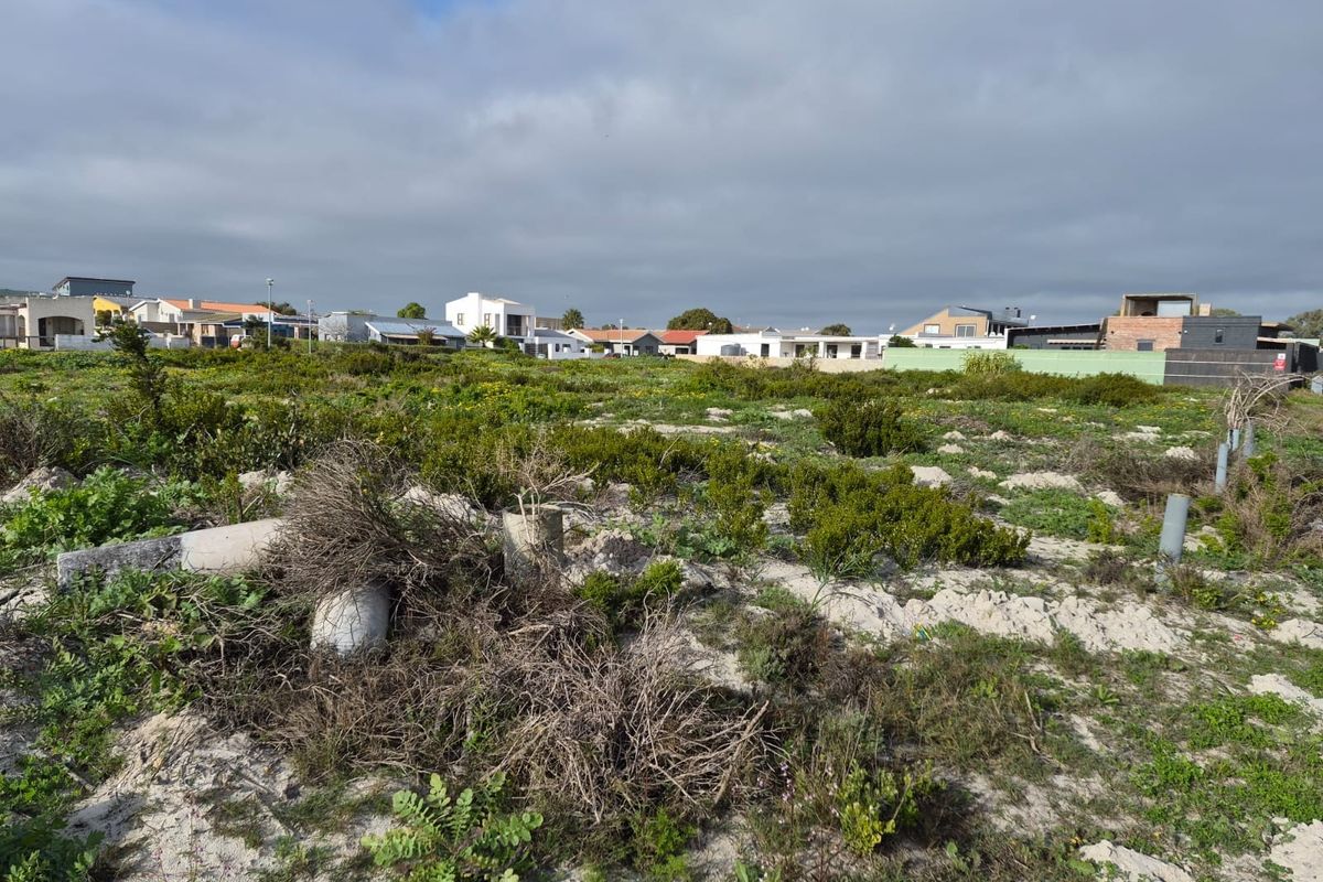 A vacant residential plot featuring natural vegetation and sandy terrain, situated in an established neighborhood with surrounding residential properties.