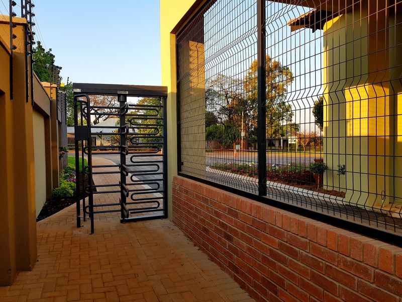 A secure pedestrian entrance featuring a metal turnstile, brick boundary wall, and mesh security fencing with integrated electric wiring.