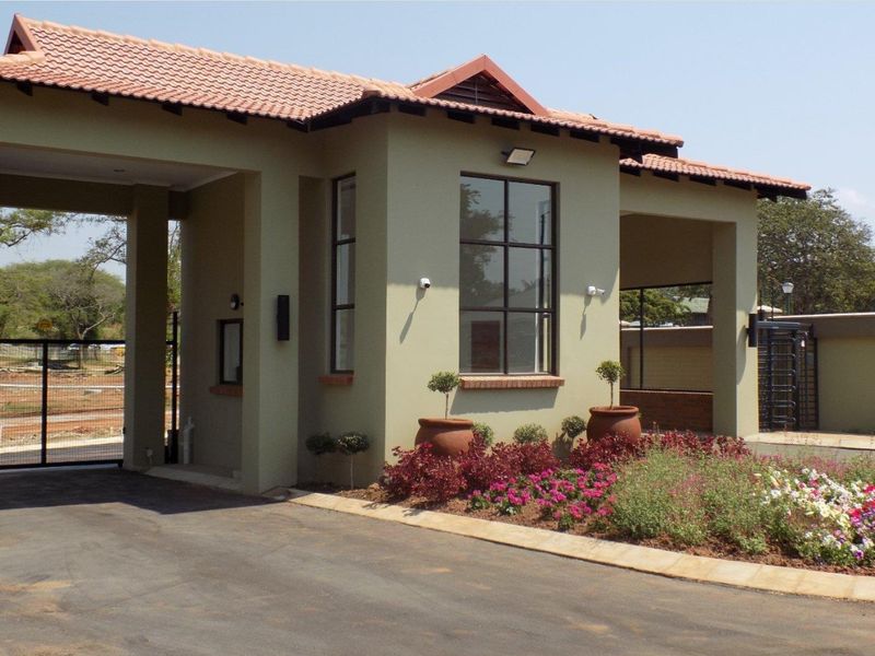A well-maintained residential security entrance gatehouse featuring a tiled roof, paved driveway, and established garden landscaping.