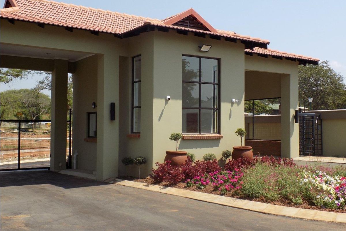 A well-maintained residential security entrance gatehouse featuring a tiled roof, paved driveway, and established garden landscaping.