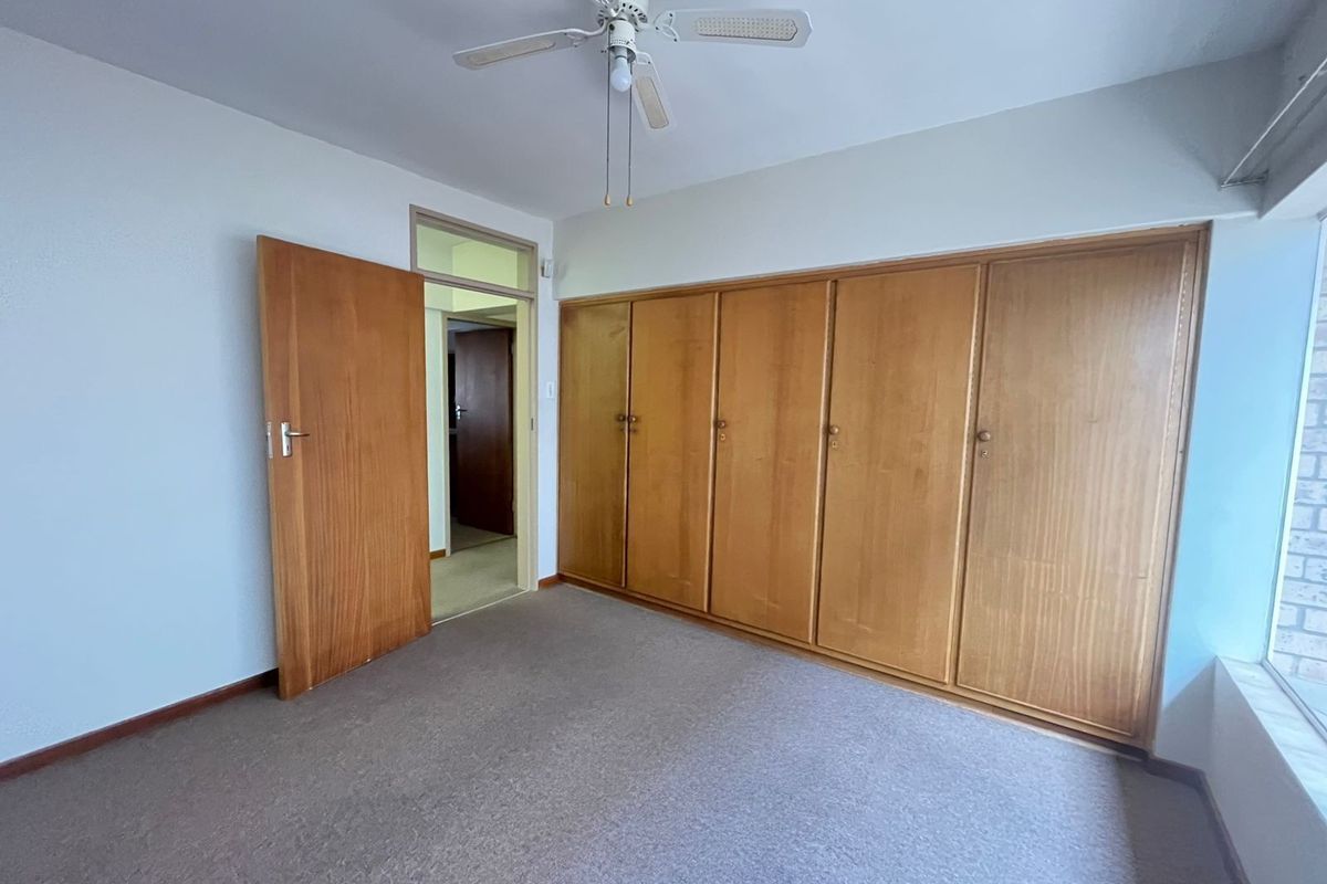 A spacious, empty bedroom featuring extensive built-in wooden cupboards, a ceiling fan, and neutral carpeting, offering a blank canvas for personalization.