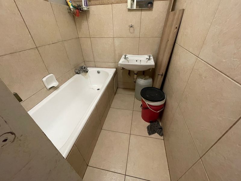A compact bathroom featuring a bathtub and wall-mounted basin with full-height ceramic tiling, currently requiring cosmetic updates and vanity installation.