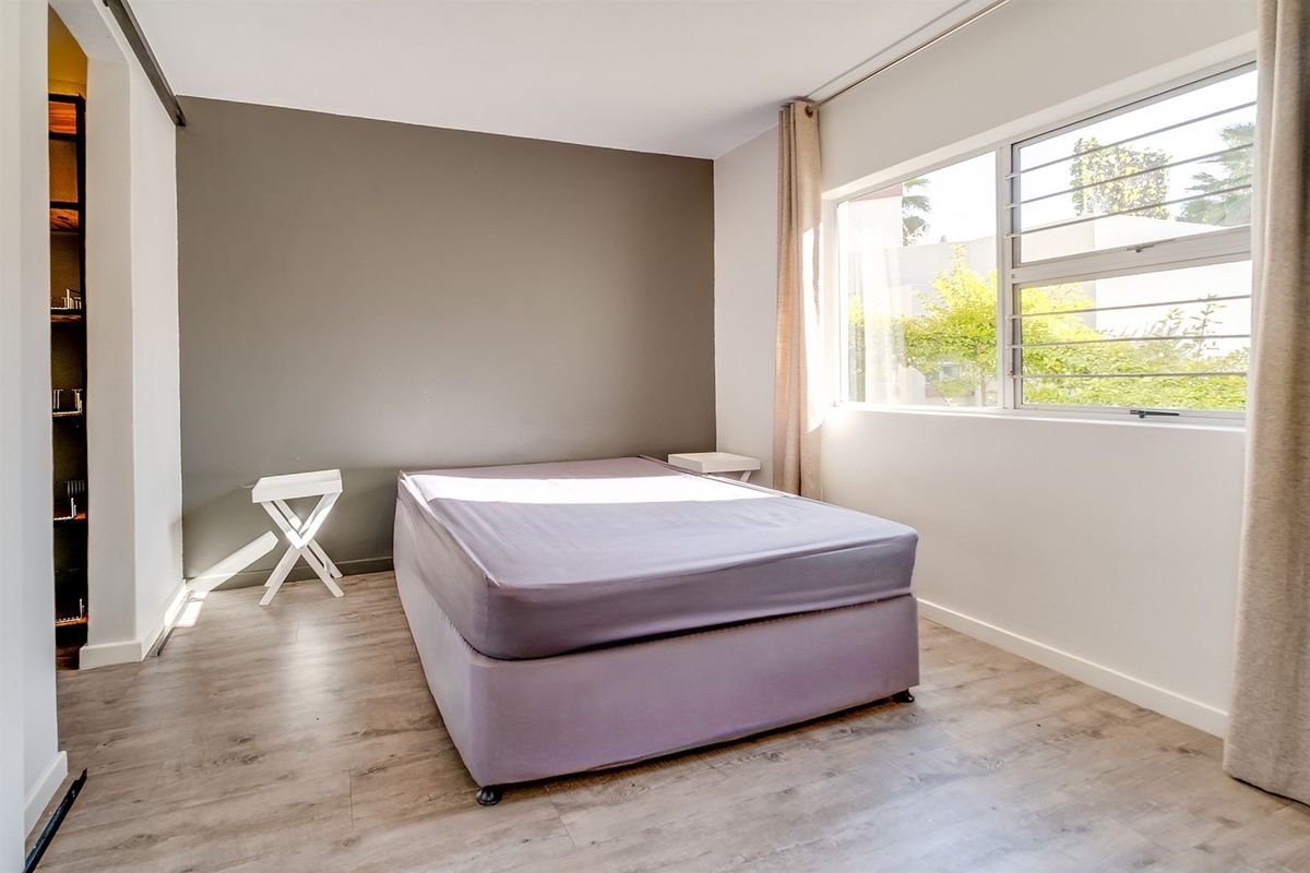 A bright bedroom featuring modern laminate flooring, a large sliding window with security bars, and a neutral grey accent wall.