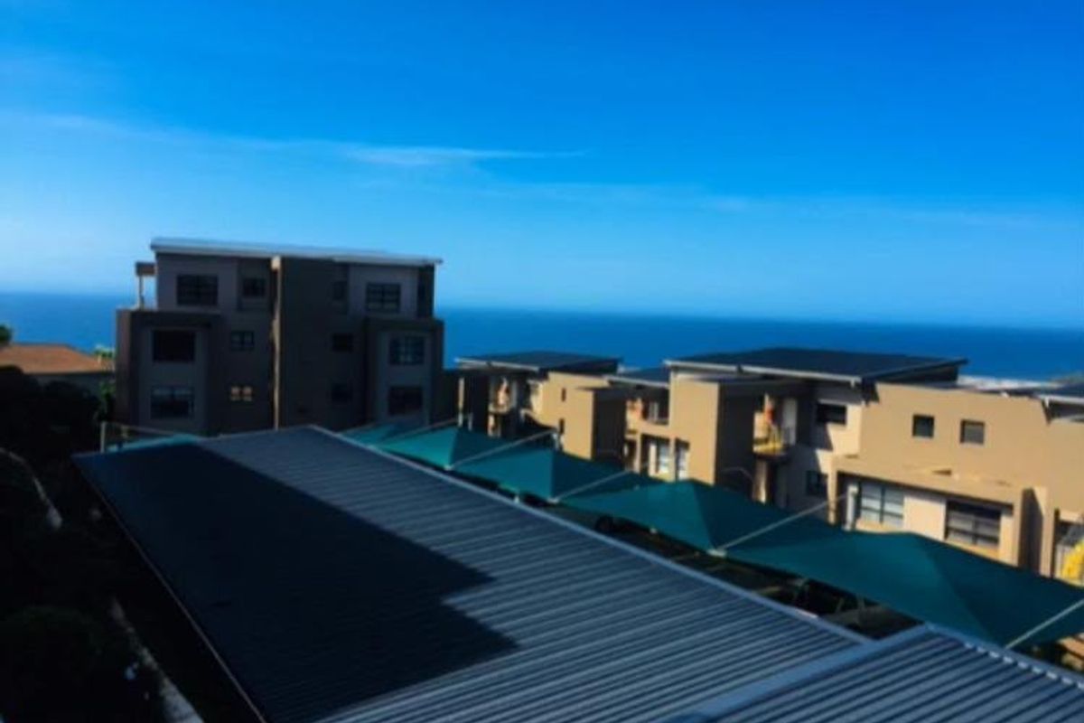 An elevated aerial view of a modern apartment complex featuring metal roofing, shade cloth parking structures, and a scenic ocean backdrop.