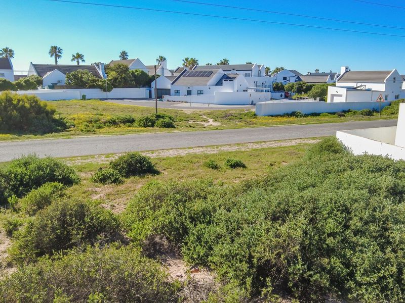 Aerial view of a residential estate featuring white-walled homes with solar panels, surrounded by natural coastal vegetation and local road infrastructure.