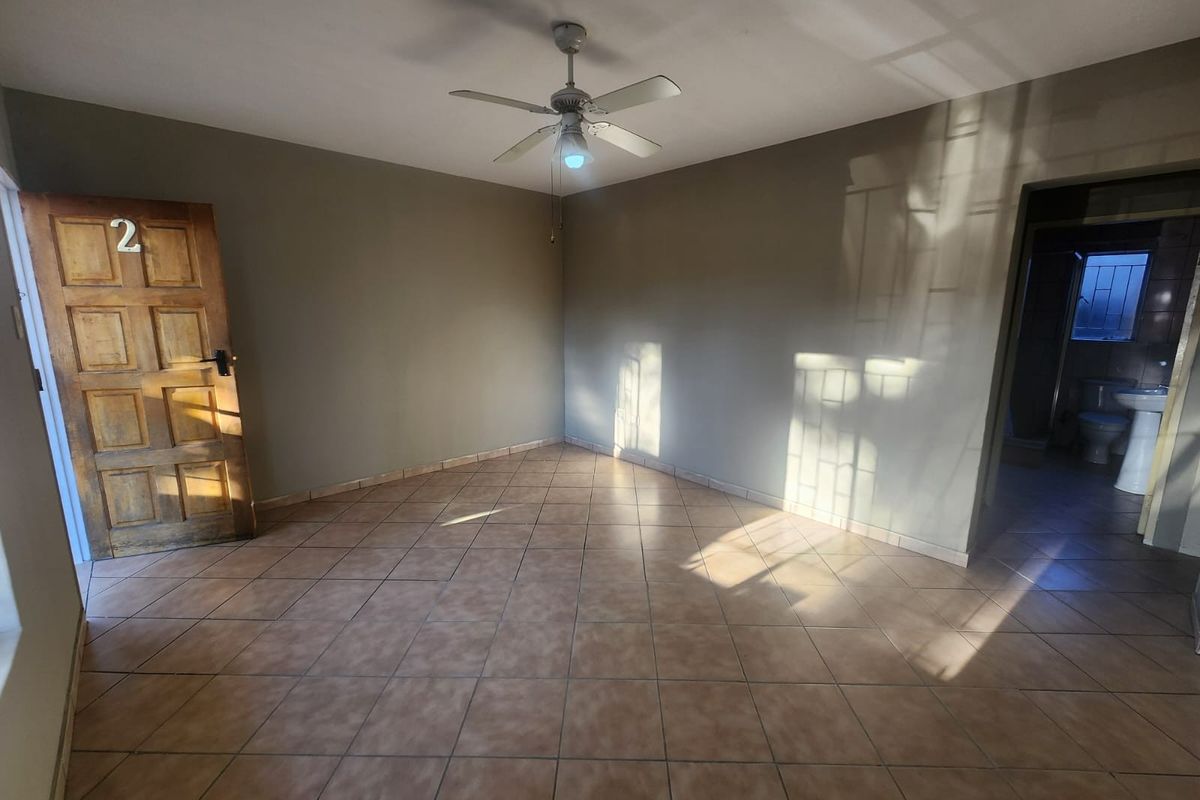 An empty bedroom featuring tiled flooring, a ceiling fan, and a wooden entrance door, with an adjacent bathroom visible.