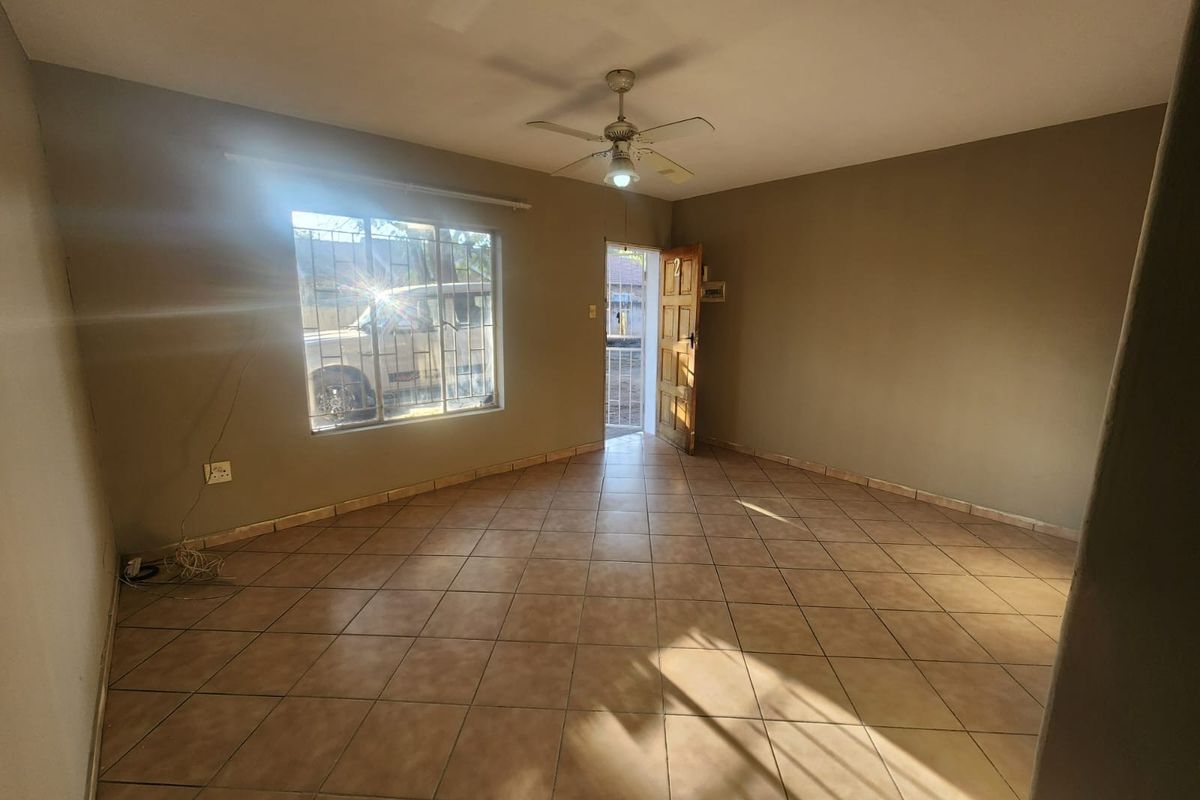 An empty living room featuring ceramic floor tiles, a ceiling fan, and a secure entrance door with natural light entering through the window.