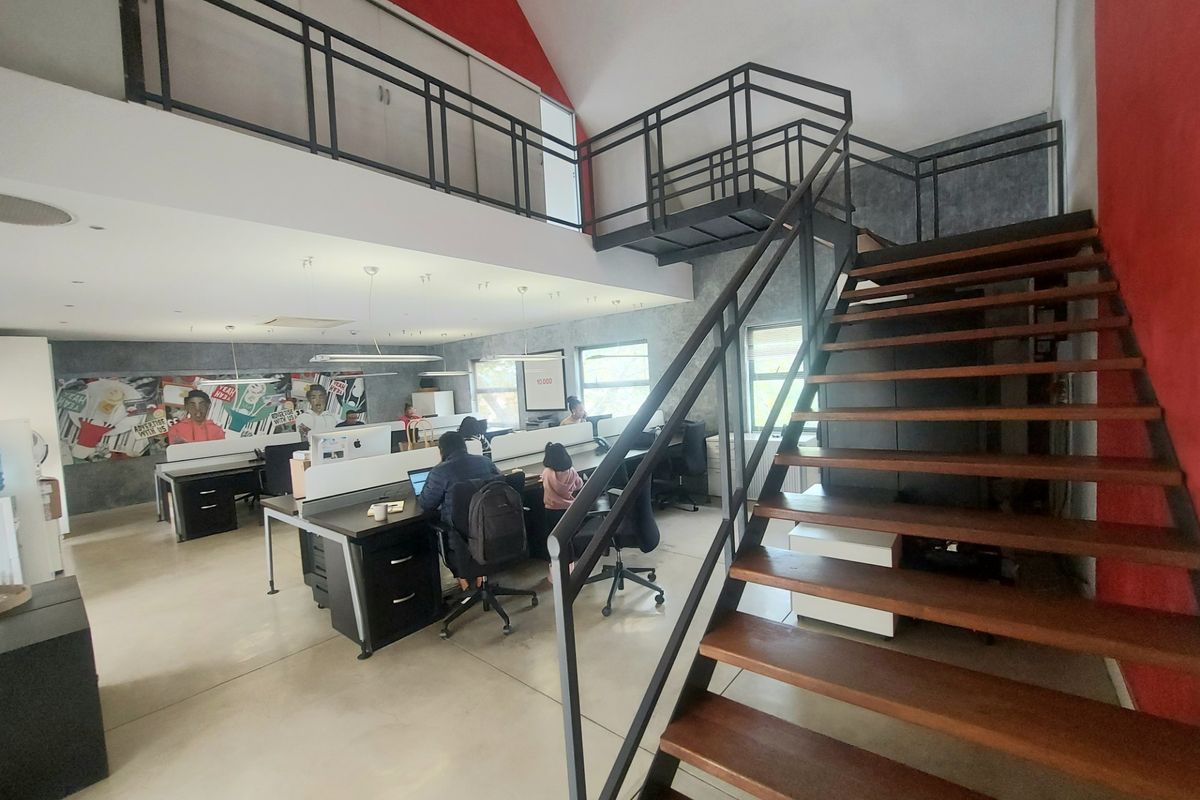 Spacious open-plan office featuring a mezzanine level, modern metal staircase, polished concrete floors, and ample natural light from large windows.