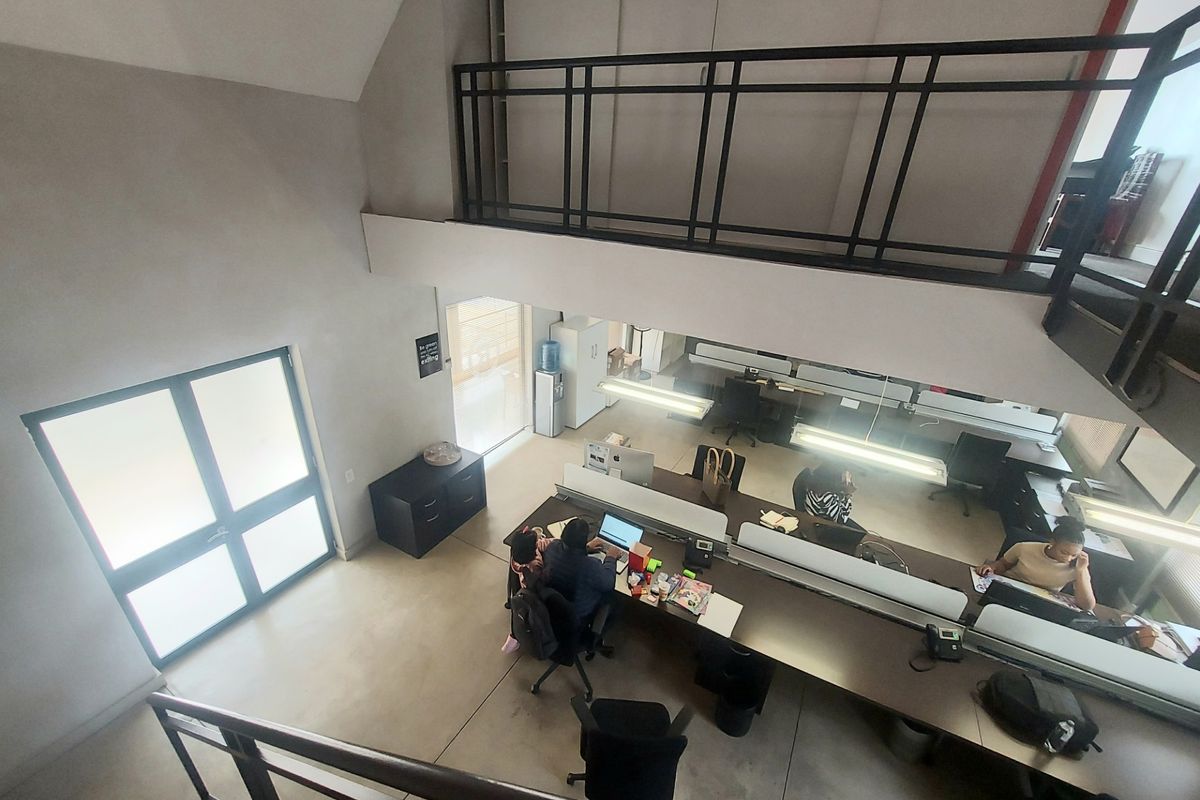 Open-plan office space featuring a mezzanine level with metal railings, built-in workstations, and ample natural light from large glass doors.