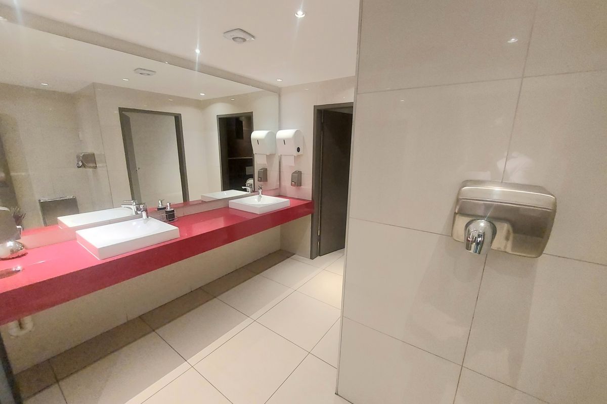Modern office bathroom featuring a double vanity with ceramic basins, large wall mirror, tiled surfaces, and integrated hand dryer.