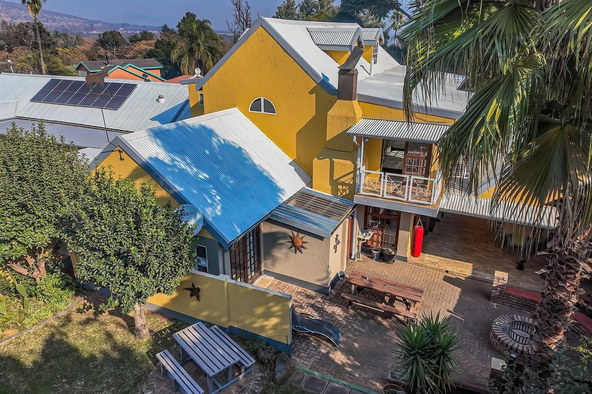 Aerial view of the rear exterior featuring a yellow facade, metal roofing, solar panels, a paved patio area, and a built-in braai.