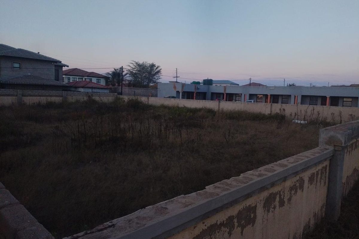 A vacant residential plot enclosed by a perimeter wall, featuring overgrown grass and clear boundaries in a suburban neighborhood setting.