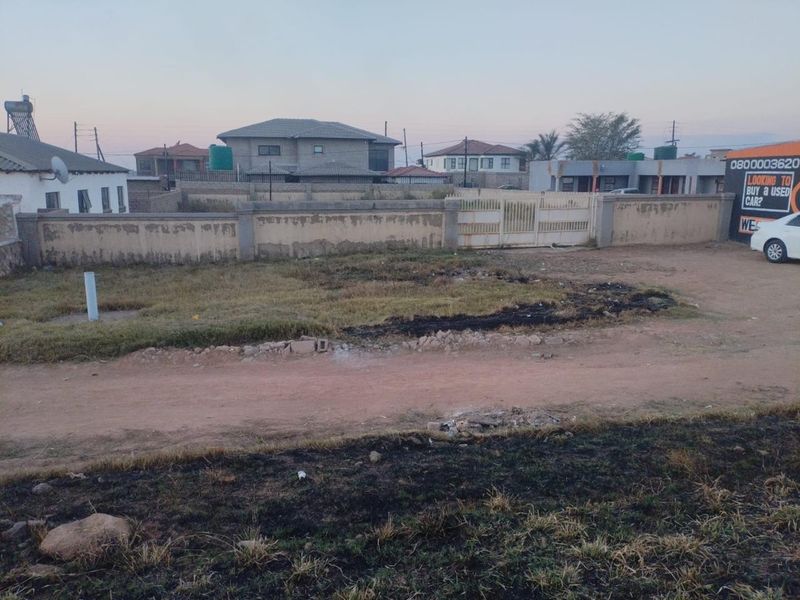 A vacant residential plot featuring a perimeter wall and metal gate, situated along an unpaved access road in a suburban neighborhood.