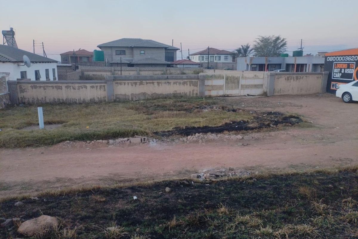 A vacant residential plot featuring a perimeter wall and metal gate, situated along an unpaved access road in a suburban neighborhood.