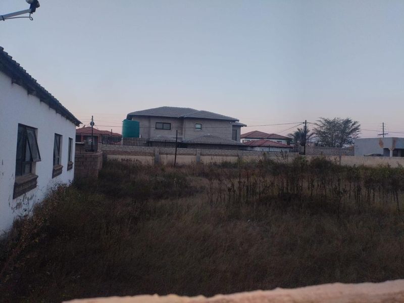 A view of a vacant residential plot featuring a perimeter wall and a water storage tank, with surrounding neighborhood properties visible.