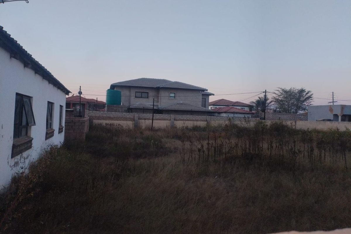 A view of a vacant residential plot featuring a perimeter wall and a water storage tank, with surrounding neighborhood properties visible.