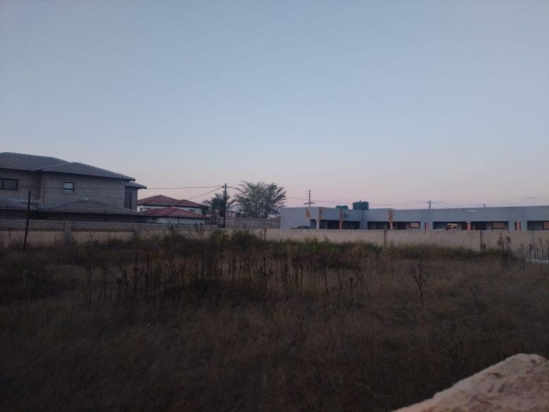 A view of a vacant residential plot featuring dry vegetation and enclosed by a perimeter wall with neighboring properties visible in the background.