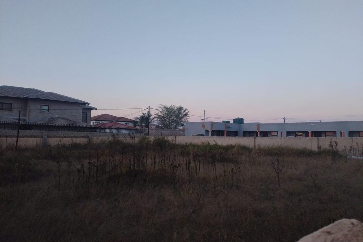 A view of a vacant residential plot featuring dry vegetation and enclosed by a perimeter wall with neighboring properties visible in the background.