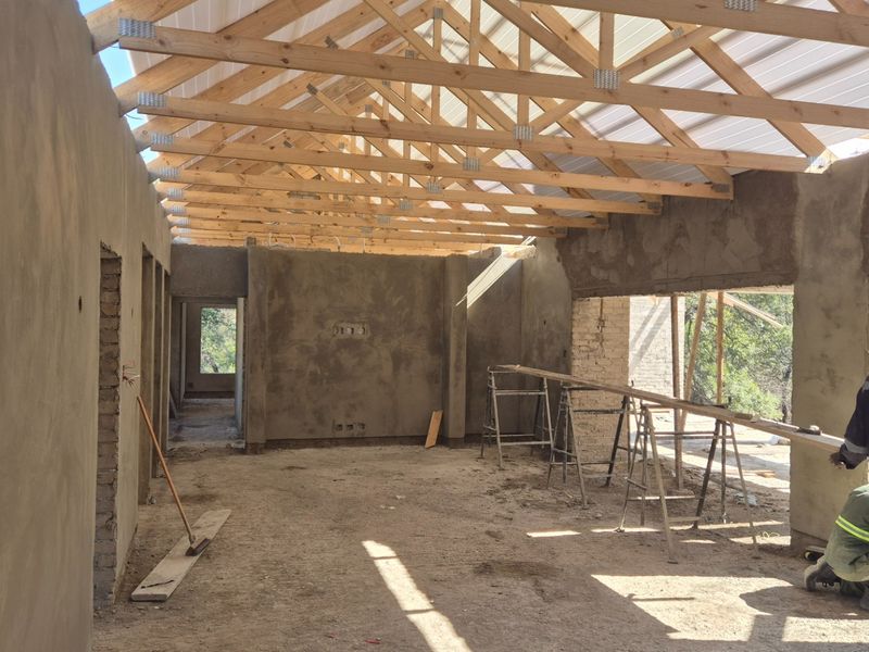 An open-plan interior space under construction, featuring exposed timber roof trusses, plastered walls, and rough-in electrical points, awaiting final finishes and flooring.