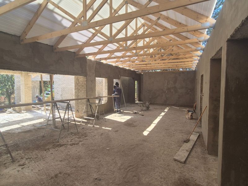 An open-plan interior space currently under construction, featuring exposed timber roof trusses, plastered brick walls, and a concrete floor.