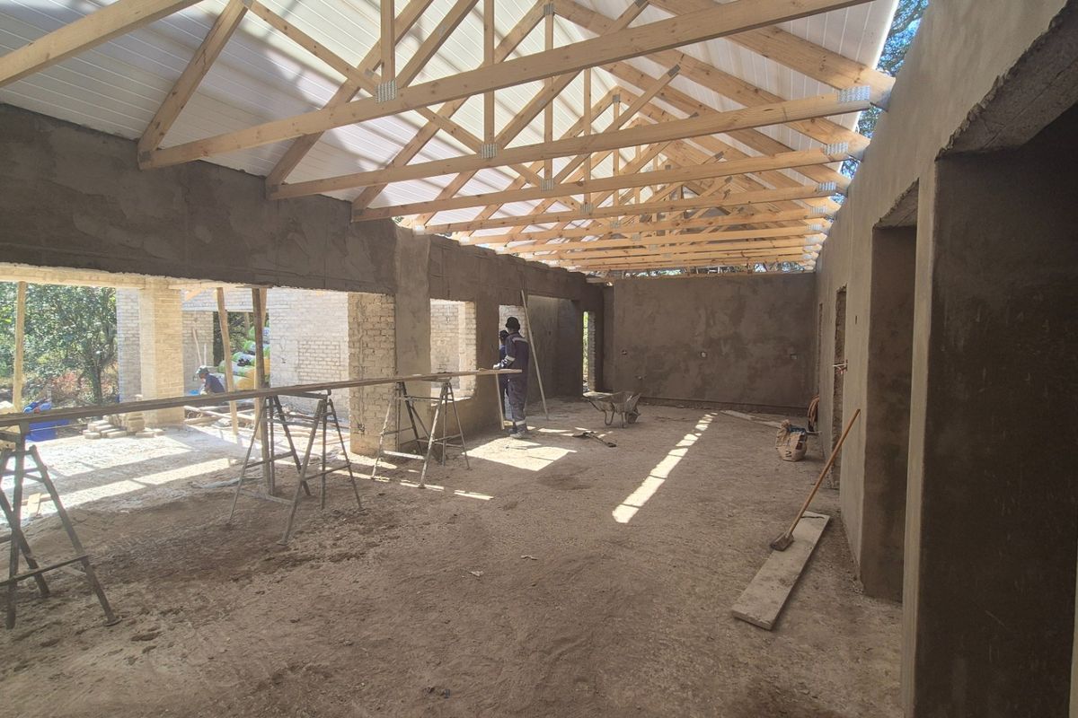 An open-plan interior space currently under construction, featuring exposed timber roof trusses, plastered brick walls, and a concrete floor.