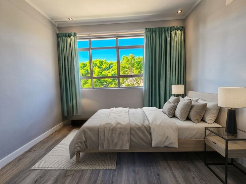 A well-lit bedroom featuring modern laminate flooring, recessed ceiling lighting, and a large window providing natural light and a pleasant outdoor view.