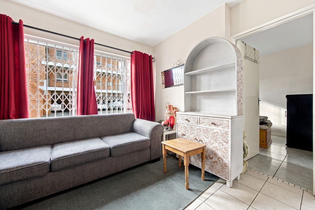 A living room area featuring tiled flooring and a large window fitted with a security gate, providing natural light and access to the hallway.