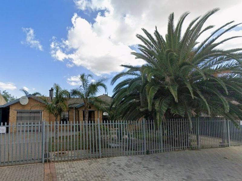 Front elevation of a single-story brick house featuring a tiled roof, attached garage, and secure metal perimeter fencing.